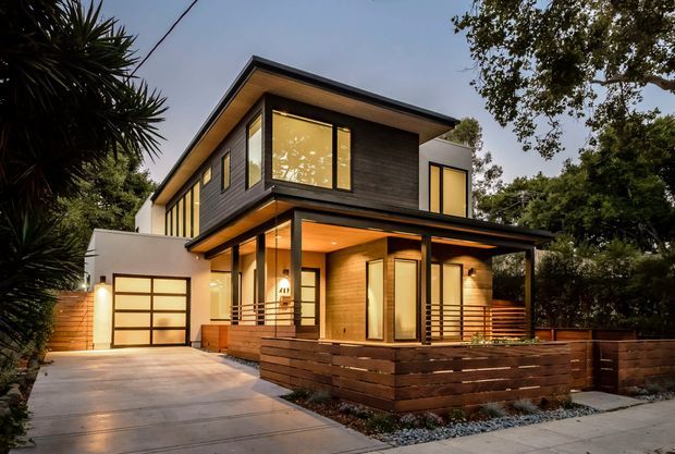A contemporary two-storey suburban house photographed at dusk, with a sharply angled low-pitched roof, dark horizontal cladding, warm timber soffits and extensive illuminated glazing. A covered timber-decked entry and porch project toward the front, alongside a single garage with a translucent panel door. The property features timber fencing, concrete driveway paving and landscaped planting around the perimeter.