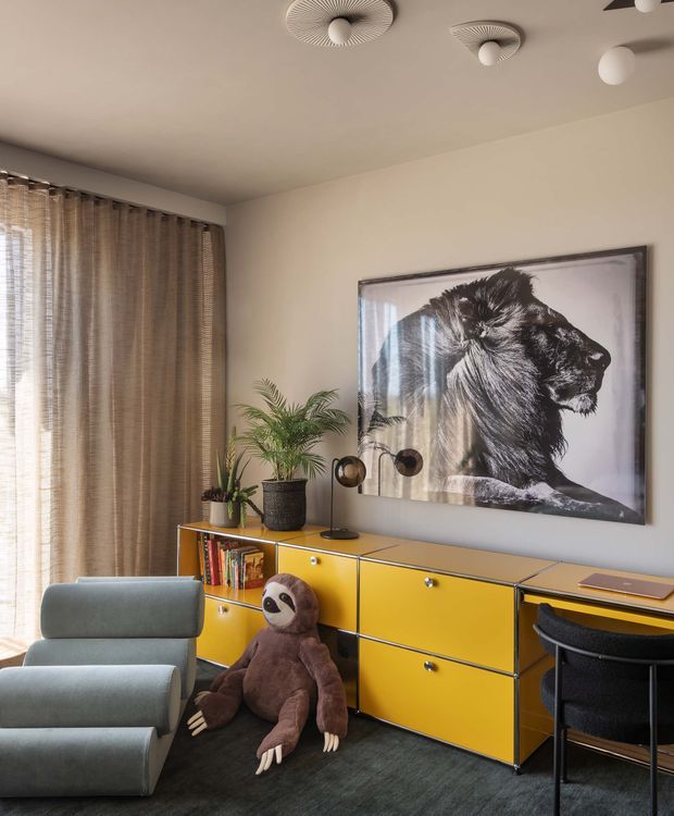 A stylish, playful interior corner featuring a long mustard-yellow built-in sideboard with open shelving and books, a large black-and-white lion artwork, potted plants, sculptural table lamps, and a desk or work surface with a dark chair. A muted blue-grey lounge chair and plush sloth toy sit on a dark carpet beside full-height textured curtains. The room has a warm neutral palette and distinctive sculptural ceiling light fixtures.