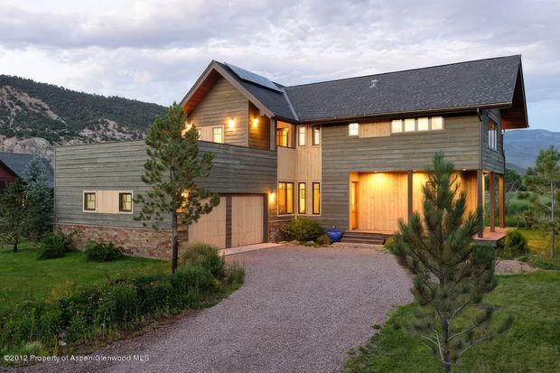 Exterior view of a large multi-level mountain home with a steep gabled roof, dark grey roof shingles, horizontal timber cladding, stonework at the lower level, and warm illuminated windows and entry doors. A gravel driveway, landscaped garden, young conifers, and mountainous terrain surround the house.