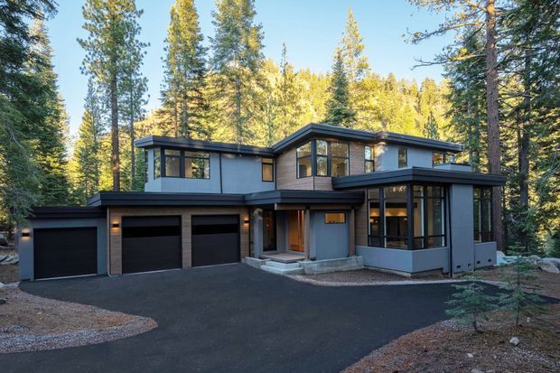A contemporary multi-level house nestled among tall forest trees. The dark grey and black exterior features broad glazing, warm timber accents, a low-pitched roof, projecting volumes, and an attached three-car garage. A paved driveway and gravel-edged landscaping lead to the recessed entry.