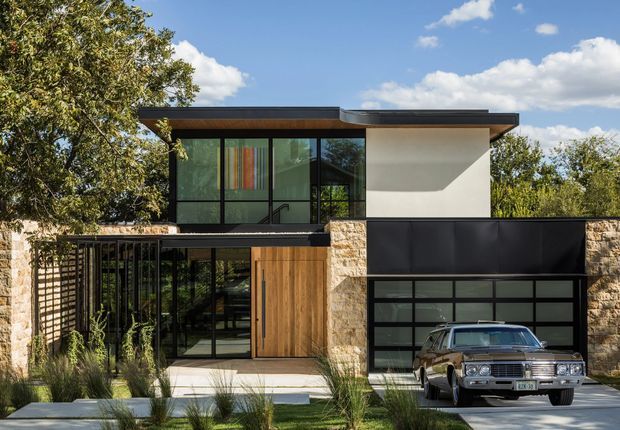 A contemporary two-storey residential facade set among mature trees, combining dark metal-framed glazing, pale stone masonry, smooth white rendered walls, warm vertical timber cladding, and broad low-pitched roof planes. A black-framed garage occupies the lower right, with a vintage car parked in the driveway. The upper level includes expansive glazing and a projecting roof overhang, while ornamental grasses and concrete stepping slabs soften the entry landscape.