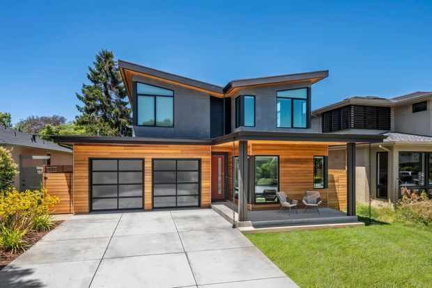 Exterior view of a contemporary two-storey suburban residence with a bold asymmetrical roofline, dark upper-level cladding, warm timber-look horizontal cladding, expansive black-framed glazing, an attached double garage, concrete driveway, and a covered ground-floor patio.