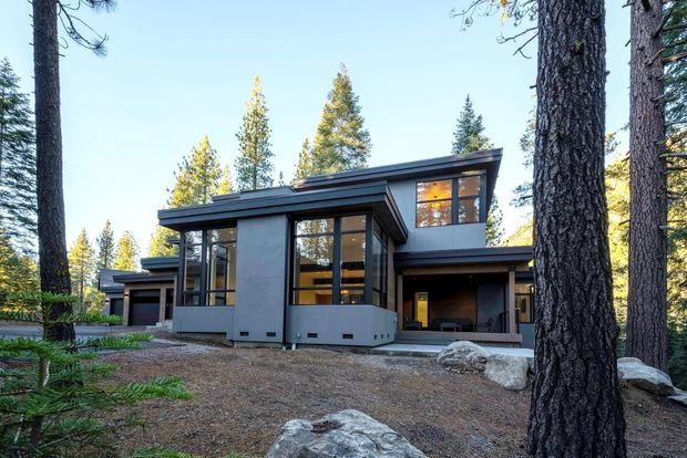 A modern multi-level house set among tall pine trees in a forested setting. The building features flat and low-pitched roof planes, dark horizontal cladding, expansive floor-to-ceiling glazing, projecting window bays, and a sheltered recessed entrance or outdoor living area. Natural rock and wooded terrain surround the home.
