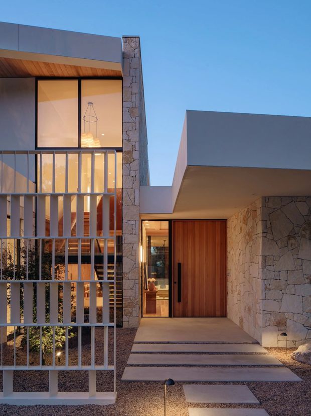 Contemporary residential facade photographed at dusk, featuring a flat-roofed composition with projecting geometric volumes, extensive floor-to-ceiling glazing, a warm timber soffit and front door, pale irregular stone-clad walls, and a vertical metal privacy screen enclosing the glazed entry-side facade. Stepping-stone paving crosses a gravel forecourt, while warm interior lighting creates an inviting glow.