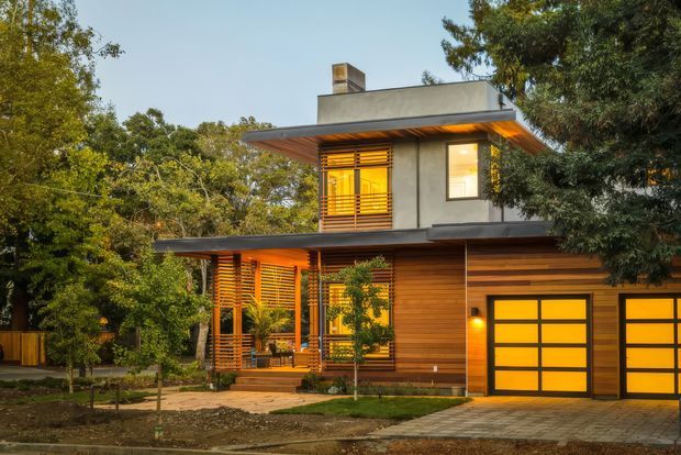 Exterior view of a contemporary two-storey residence set among mature trees. The house combines horizontal timber cladding and timber privacy screens with smooth light rendered walls, broad flat roof overhangs, and large glazed openings. A double garage with illuminated frosted or translucent-panel doors occupies the lower right, while warm interior lighting creates an inviting evening atmosphere.
