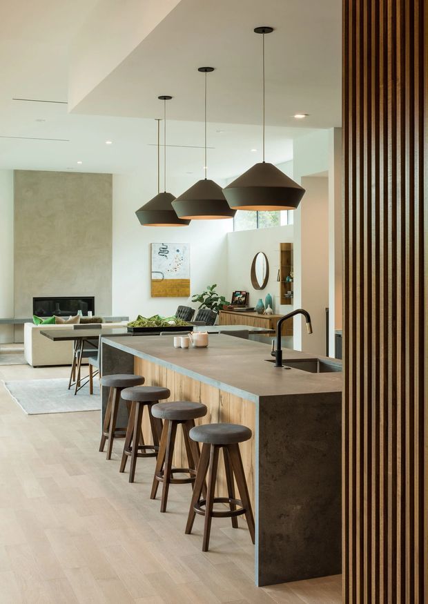 Open-plan contemporary kitchen and living area with a large island, integrated sink and black tap, timber-fronted cabinetry, dark stone-look benchtop, and four bar stools. Three oversized dark pendant lights hang above the island. A living room with fireplace and built-in seating is visible beyond, while a vertical timber slat screen frames the right side. The space has pale timber flooring, white walls and ceiling, and a calm, refined neutral palette.