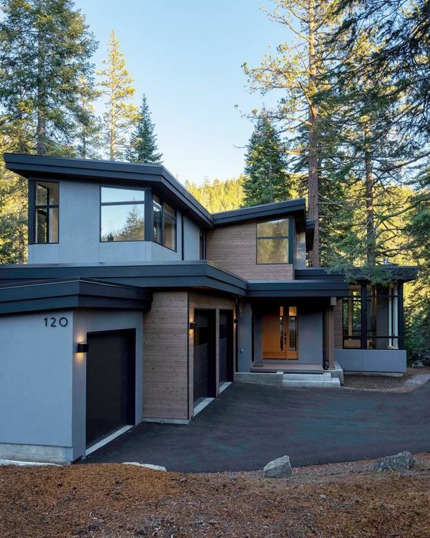 Exterior photograph of a contemporary multi-level residence set among mature forest trees. The house features flat and low-pitched roof planes, dark metal trim, grey rendered walls, warm timber cladding, extensive black-framed glazing, a recessed timber entry door, and an integrated single garage. A paved driveway and low rock edging occupy the foreground.