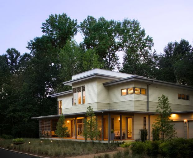 Two-storey contemporary residence set among mature trees, with pale horizontal cladding, broad low-pitched roof planes, projecting eaves, warm timber-framed glazing, and illuminated interiors. The landscaped foreground and evening lighting give the house a calm, natural suburban or bush setting.
