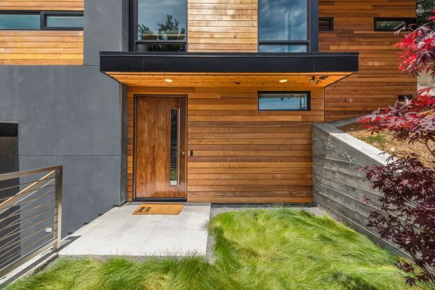 Contemporary residential front entry with warm horizontal timber cladding, dark grey rendered or concrete wall panels, tall glazed windows, and a substantial timber pivot-style door. A black projecting canopy shelters the entrance, while a steel-and-timber balustrade, concrete path, retaining wall, ornamental planting, and ornamental grasses complete the compact landscaped approach.