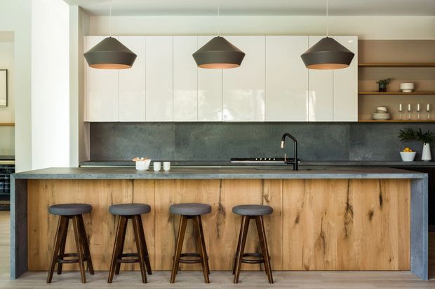 Contemporary kitchen centred on a large island with a natural timber panelled base, dark stone benchtop, black sink and tapware, glossy white full-height cabinetry, and a dark textured splashback. Four timber stools sit beneath three oversized dark pendant lights, while open timber shelving with crockery and plants is visible at right.