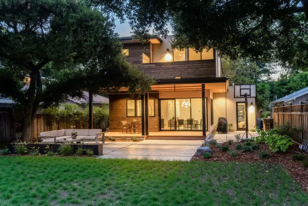 Twilight rear exterior view of a contemporary two-storey house opening onto a landscaped backyard. The home features warm horizontal timber cladding, extensive glazing, a covered patio, outdoor seating, paved terrace, garden beds, and a basketball hoop.