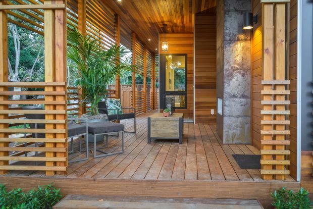A covered timber outdoor veranda or entry terrace surrounded by lush vegetation. The space features warm timber decking, ceiling and slatted privacy screens, modern outdoor lounge seating, a small coffee table, potted tropical plants, a large natural-stone-clad structural column, and a glazed entry door. The design creates a warm, sheltered connection between the home and garden.