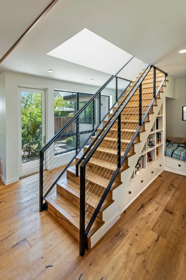Bright contemporary stairway within an open residential interior. The staircase features warm timber treads and stringers, black metal handrails and posts, and horizontal wire balustrading. White built-in cabinetry and shelving are integrated beneath and beside the stairs, with a small built-in reading nook. A large skylight, recessed ceiling lights, timber flooring, and full-height glazed doors create an airy, well-lit space.