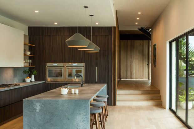 A contemporary open-plan kitchen with a large concrete island and integrated seating, dark timber cabinetry, built-in double ovens, open shelving, and pendant lights. A short set of timber steps leads to a raised hallway or adjacent space, while full-height glazing brings in views of lush greenery. The space has a warm, refined material palette with strong indoor-outdoor connection.