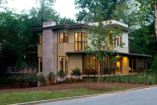 A contemporary two-storey house set within a wooded, landscaped site. The exterior combines horizontal light-toned cladding, a prominent tall stone-clad chimney, broad roof overhangs, extensive timber louvre screens, and warm interior lighting visible through the windows. A covered outdoor area extends along the right side of the home.