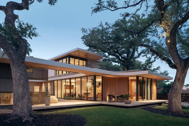 A sophisticated multi-level contemporary residence nestled among mature oak trees. The home features extensive floor-to-ceiling glazing, warm timber-lined walls and soffits, slender dark frames, cantilevered roof planes, and gently curving roof edges surrounding a broad outdoor terrace. The dusk setting highlights the warm interior lighting and strong connection between the living spaces and landscaped garden.