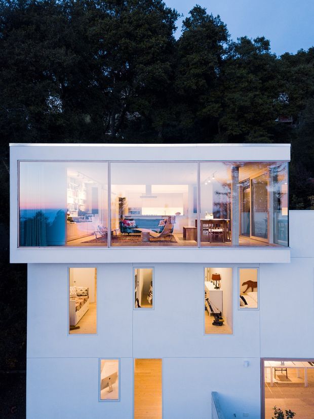 Twilight exterior view of a minimalist modern multi-level home set against dense trees. The white cubic facade has extensive floor-to-ceiling glazing on the upper level, revealing a bright open-plan living area, kitchen, dining space and timber-lined interior. Smaller windows and a timber entry door punctuate the lower levels, with a partial courtyard or terrace visible at the base.