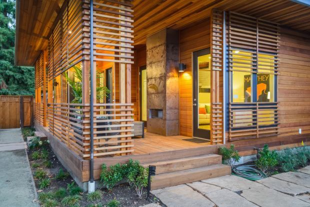 Exterior view of a warm contemporary timber-clad house with a covered entry porch, horizontal timber privacy screens, vertical steel posts, timber decking and steps, stone feature wall, and illuminated interior visible through the entry door and window. Landscaping, paving and a garden fence surround the facade.