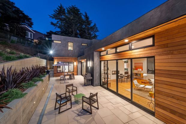 Twilight view of a modern courtyard-style residence with a low, flat-roofed form, dark concrete walls, warm horizontal timber cladding, and extensive floor-to-ceiling glazing. A paved outdoor terrace contains casual seating and a barbecue, while the illuminated interior reveals a dining area and contemporary furnishings. Integrated planters and a stepped sloping site frame the courtyard.