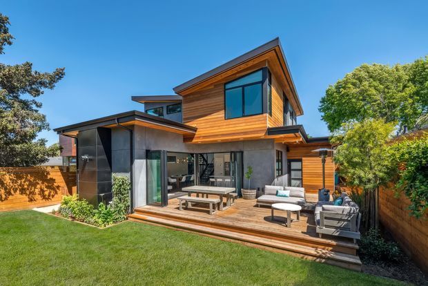 A contemporary two-storey residence viewed from a landscaped backyard, featuring warm horizontal timber cladding, dark metal accents, broad glazing, and a covered timber deck with outdoor lounge and dining furniture. The house has offset roof forms and strong indoor-outdoor connection, opening onto a manicured lawn enclosed by timber fencing and mature planting.
