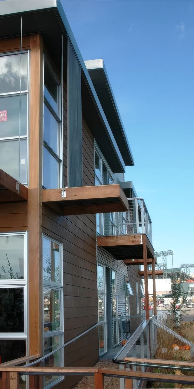 Exterior architectural photograph of a contemporary multi-storey building with dark horizontal cladding, extensive glazing, exposed timber balcony structures, metal railings, and projecting roof overhangs. The image looks along an elevated access walkway or balcony toward an urban streetscape under a clear blue sky.