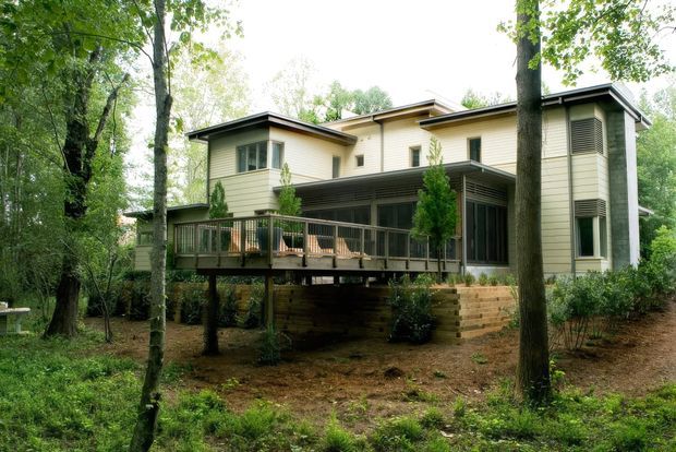 A contemporary multi-level house set among mature trees, viewed from the exterior. The home features pale horizontal weatherboard cladding, dark low-pitched roof sections, extensive screened glazing, and an elevated timber deck supported above the wooded ground. Layered timber retaining walls and landscaped planting sit beneath and around the house.