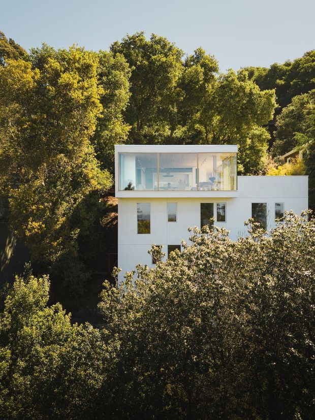 Exterior view of a modern white multi-storey house set into a densely wooded, sloping site. The upper level is defined by a projecting glass-walled room with slim light-coloured frames, while the lower facade has simple white walls and rectangular windows. Trees heavily surround and partially obscure the building.