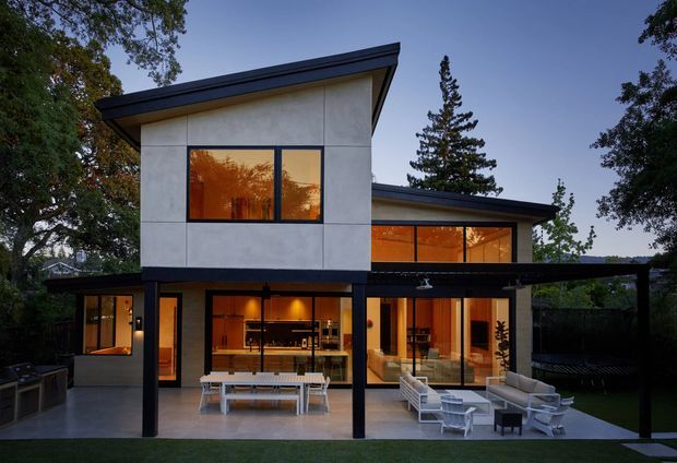 Contemporary two-storey house exterior viewed at dusk, with crisp geometric volumes, white rendered wall panels, dark flat or low-pitched roof planes, and extensive warm-lit glazing. A covered outdoor terrace with lounge and dining furniture sits in front of the living areas, surrounded by mature trees and landscaped grounds.
