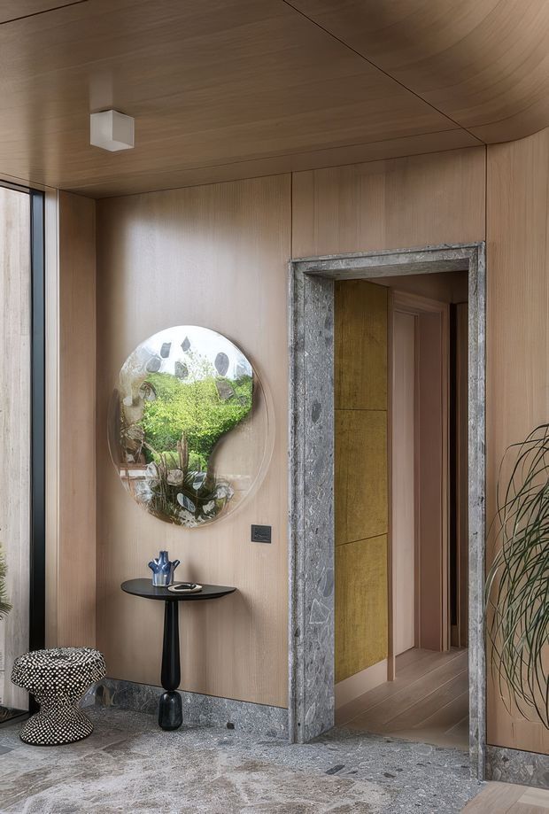 A refined interior entry or hallway with warm timber wall and ceiling panelling, a curved ceiling edge, and a terrazzo-like grey stone floor and skirting. A rough natural-stone doorway frames a passage into adjoining rooms. The space includes a sculptural wall mirror, slim black console table, decorative objects, patterned stool, and indoor planting. Floor-to-ceiling glazing is visible at left, bringing in daylight. The overall design is tactile, contemporary, and luxurious with natural materials and sculptural detailing.