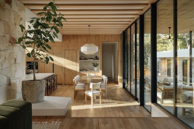 Warm contemporary dining and living space with a timber-lined ceiling and walls, exposed ceiling beams, broad timber flooring, and full-height black-framed glazing along one side. A wooden dining table with sculptural white chairs sits beneath a faceted pendant light, while a stone-clad feature wall, built-in shelving, and potted tree add natural texture. The glazing opens visually to an adjoining outdoor or courtyard area.