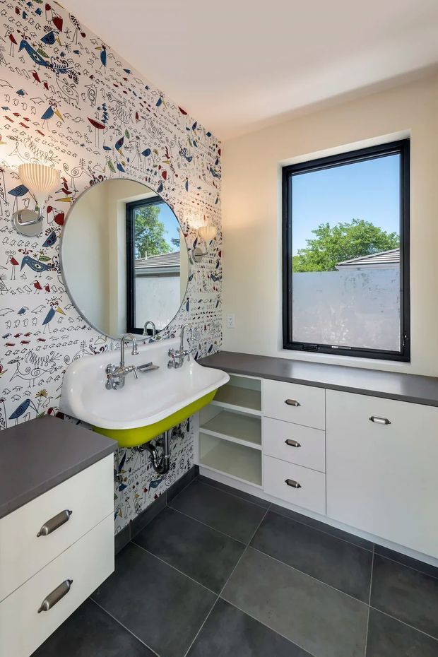 A bright, playful bathroom featuring a wall-mounted white ceramic basin with a vivid lime-green underside, chrome taps, an oval mirror, and white built-in cabinetry with dark benchtops. The focal wall is covered in whimsical illustrated wallpaper, while the floor uses large dark grey tiles. A black-framed window brings in natural light.