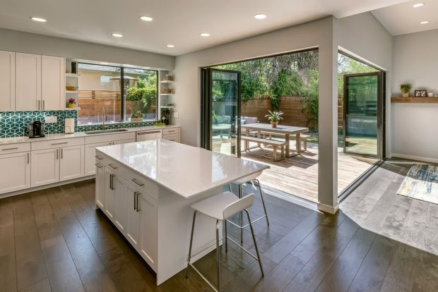 Bright, open-plan contemporary kitchen with a large white island and integrated seating, white shaker-style cabinetry, and a patterned teal geometric tile splashback. Dark timber flooring extends toward expansive black-framed glazed doors opening onto a timber deck and landscaped courtyard. Recessed ceiling lighting and minimal open shelving create a clean, indoor-outdoor living connection.
