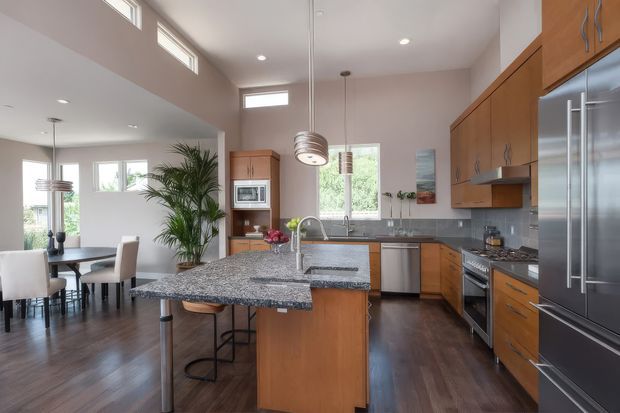 Bright open-plan kitchen and dining area with a large granite island, warm timber cabinetry, stainless-steel appliances, dark timber flooring, and high clerestory windows. A dining table and chairs are visible to the left, while pendant lights and recessed ceiling lighting illuminate the space.