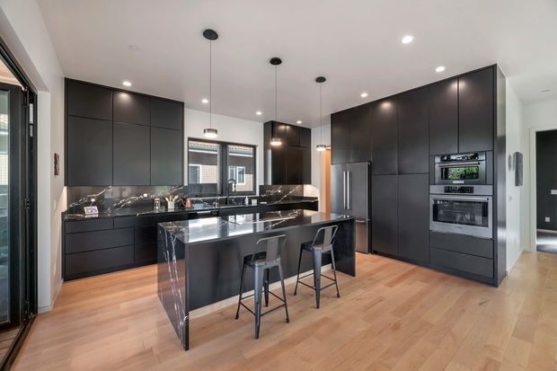 A spacious contemporary kitchen with full-height matte dark cabinetry, integrated stainless-steel appliances, a large central island with a dramatic dark marble or stone waterfall benchtop, and two bar stools. Warm timber flooring contrasts with the black cabinetry, while pendant lights and recessed ceiling lighting illuminate the room. A glazed door and windows provide natural light.
