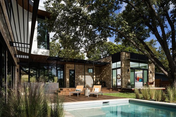 Exterior view of a low-slung contemporary residence arranged around a landscaped pool terrace. The house combines extensive floor-to-ceiling glazing, dark metal framing, warm timber soffits and decking, and rough natural stone walls beneath mature trees. A broad outdoor deck with lounge chairs overlooks the swimming pool, with ornamental grasses forming the foreground landscaping.