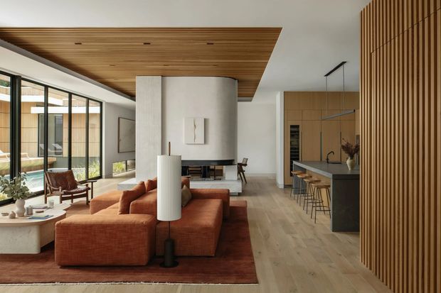 A spacious open-plan contemporary living area with a warm, sculptural interior. A rust-coloured sectional sofa and matching rug face a curved plaster fireplace feature, while full-height glazing opens toward an exterior courtyard. The room connects directly to a streamlined kitchen with timber cabinetry, a dark island, and bar stools. A timber-lined ceiling and vertical timber slatted wall add warmth and texture, complemented by pale timber flooring and restrained furnishings.