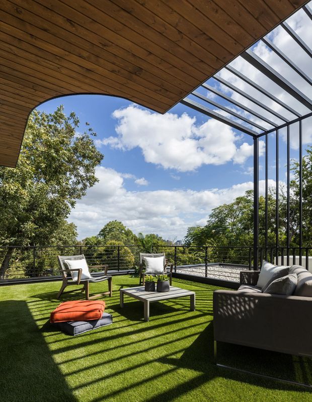 A contemporary covered outdoor terrace with a timber-lined ceiling, black metal railings, and a transparent glazed roof and side wall. The terrace is furnished as an outdoor lounge with a sofa, two armchairs, a low coffee table, cushions, and an ottoman on a green grass-like surface. Strong linear shadows from the glazing create a patterned effect, while mature trees and blue sky are visible beyond the balustrade.