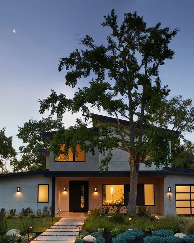 Twilight exterior view of a contemporary two-storey residence partially screened by mature trees. The home has a low-pitched dark roof, smooth light-grey rendered walls, warm timber-toned or brick-like entry cladding, large illuminated windows, a dark front door, and a projecting single-storey garage wing. A paved path crosses a landscaped front garden with ornamental grasses, shrubs, rocks, and low planting.