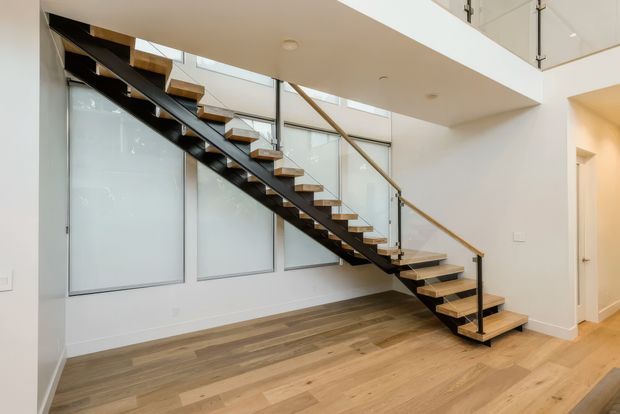 Bright contemporary interior featuring an open-riser staircase with black steel stringers, timber treads, and a glass balustrade with timber handrail. The staircase rises beside large frosted-glass panels and opens toward an upper-level void. Warm timber flooring spans the surrounding hallway or landing, with white walls and recessed ceiling lighting.