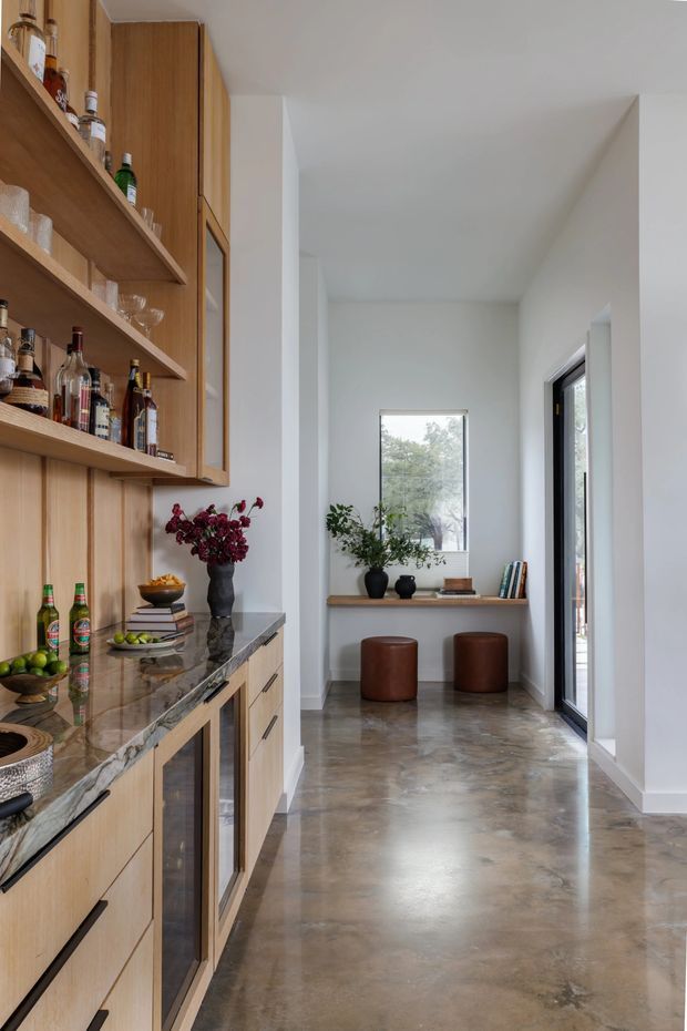 A bright, narrow residential bar and hallway space. The left side features built-in warm timber cabinetry with open shelving displaying bottles and glassware, along with a dark polished stone benchtop and integrated storage. The polished concrete floor extends toward a small seating nook with two round brown leather stools, a timber ledge, potted plants, and a window. White walls, high ceilings, and a tall glazed door create an airy, contemporary interior.