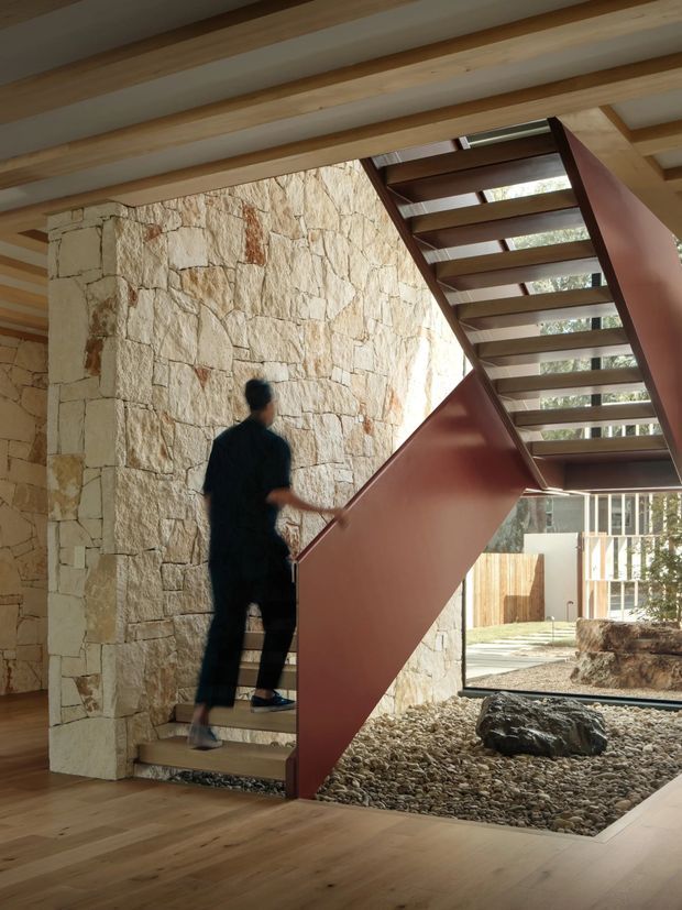 Interior view of a contemporary stairway featuring open timber treads, a sculptural rust-red metal balustrade and support, exposed light timber ceiling beams, natural stone masonry walls, and a pebble-filled indoor landscape bed. Glazing and an outdoor courtyard are visible beyond the stairs.