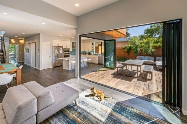 Bright open-plan interior looking from a living area toward a contemporary kitchen and dining space, with expansive black-framed sliding glass doors opening onto a timber deck and enclosed courtyard. The room features a light upholstered sectional sofa, patterned area rug, dark timber-look flooring, white cabinetry, recessed lighting, and a large indoor-outdoor connection. A cat is visible in the foreground.