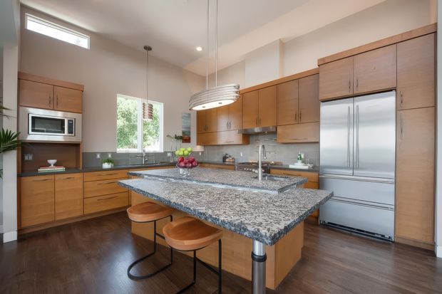 A spacious contemporary kitchen with warm wood cabinetry, dark timber flooring, stainless-steel appliances, and a large granite island with integrated sink and seating for two. The room has high ceilings, abundant natural light, recessed lighting, and a linear pendant above the island.