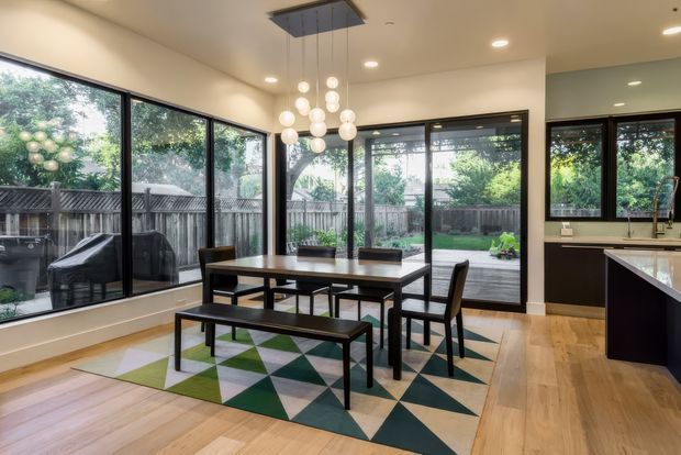 A bright, contemporary open-plan dining and kitchen area with extensive black-framed glazing overlooking a fenced garden. A dark timber dining table with bench seating sits on a geometric green, cream and charcoal rug, beneath a cluster of spherical pendant lights. The kitchen island and dark cabinetry are visible to the right, with pale timber flooring and warm off-white walls throughout.