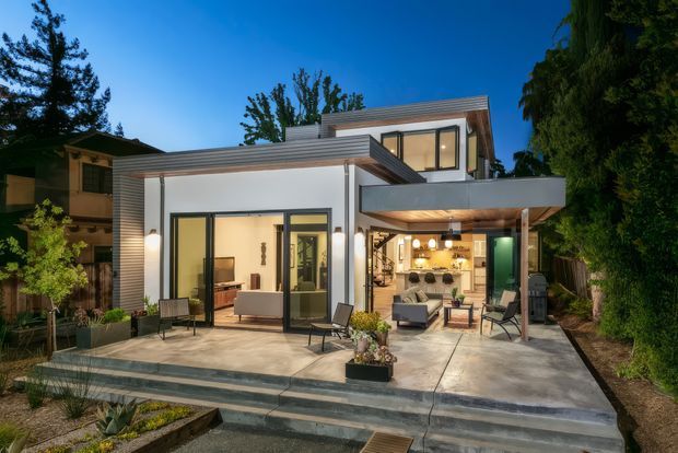Twilight exterior view of a contemporary multi-level home with a flat-roofed, cantilevered form, white rendered walls, horizontal timber-look cladding, dark-framed glazing, and a broad concrete terrace with steps. The open ground-floor living area is warmly illuminated and visible through the glazing, with outdoor seating and landscaped planting around the terrace.