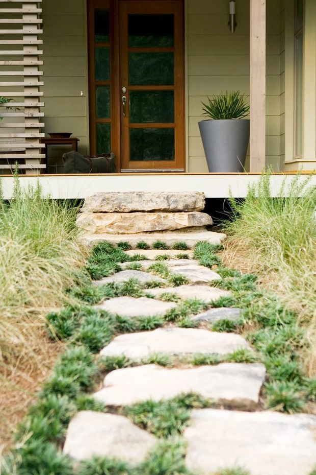 A landscaped entry path of irregular natural stone stepping slabs set through dense ornamental grass leads up to a raised front porch. The porch features pale horizontal weatherboard cladding, a timber-framed glazed entry door, a vertical timber post, a slatted timber screen, a wall light, and a large grey planter. The image focuses on the exterior approach and entry facade, with a natural, relaxed residential character.