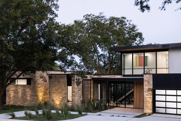 A contemporary multi-level residence viewed from the front, set among mature trees and landscaped planting. The facade combines pale natural stone walls, dark metal or painted cladding, extensive black-framed glazing, a flat roof, and a modern garage door. A glazed entry reveals a stairway and warm timber-lined interior elements, while colourful vertical artwork is visible through an upper-level window.