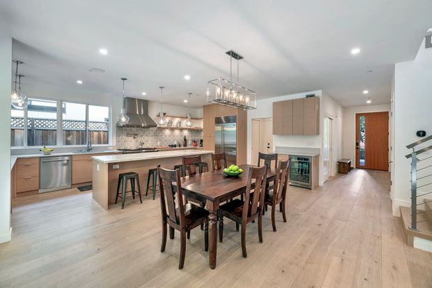 Open-plan kitchen and dining area with light timber flooring, warm wood cabinetry, a large island with bar seating, stainless-steel appliances, tiled splashback, and a dark timber dining table beneath a rectangular pendant light. The space opens toward an entry hallway and stairway, with bright recessed lighting and abundant glazing.