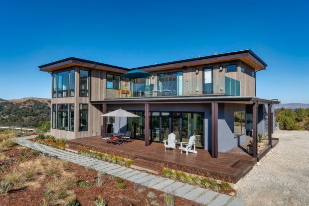 Exterior view of a contemporary two-storey home on a landscaped, sloping site. The house features extensive floor-to-ceiling glazing, warm vertical timber cladding, dark structural posts and trim, broad low-pitched roof planes, upper-level balconies, and a large timber deck with outdoor seating.