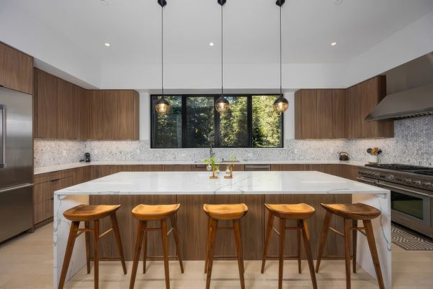 A spacious contemporary kitchen with a large white waterfall-edge island and five wooden bar stools. Warm wood-grain cabinetry runs along the walls and island, complemented by a light speckled stone backsplash and matching pale flooring. A dark-framed horizontal window brings in views of greenery, while three pendant lights hang over the island. Stainless-steel appliances and a broad range hood complete the polished, inviting space.